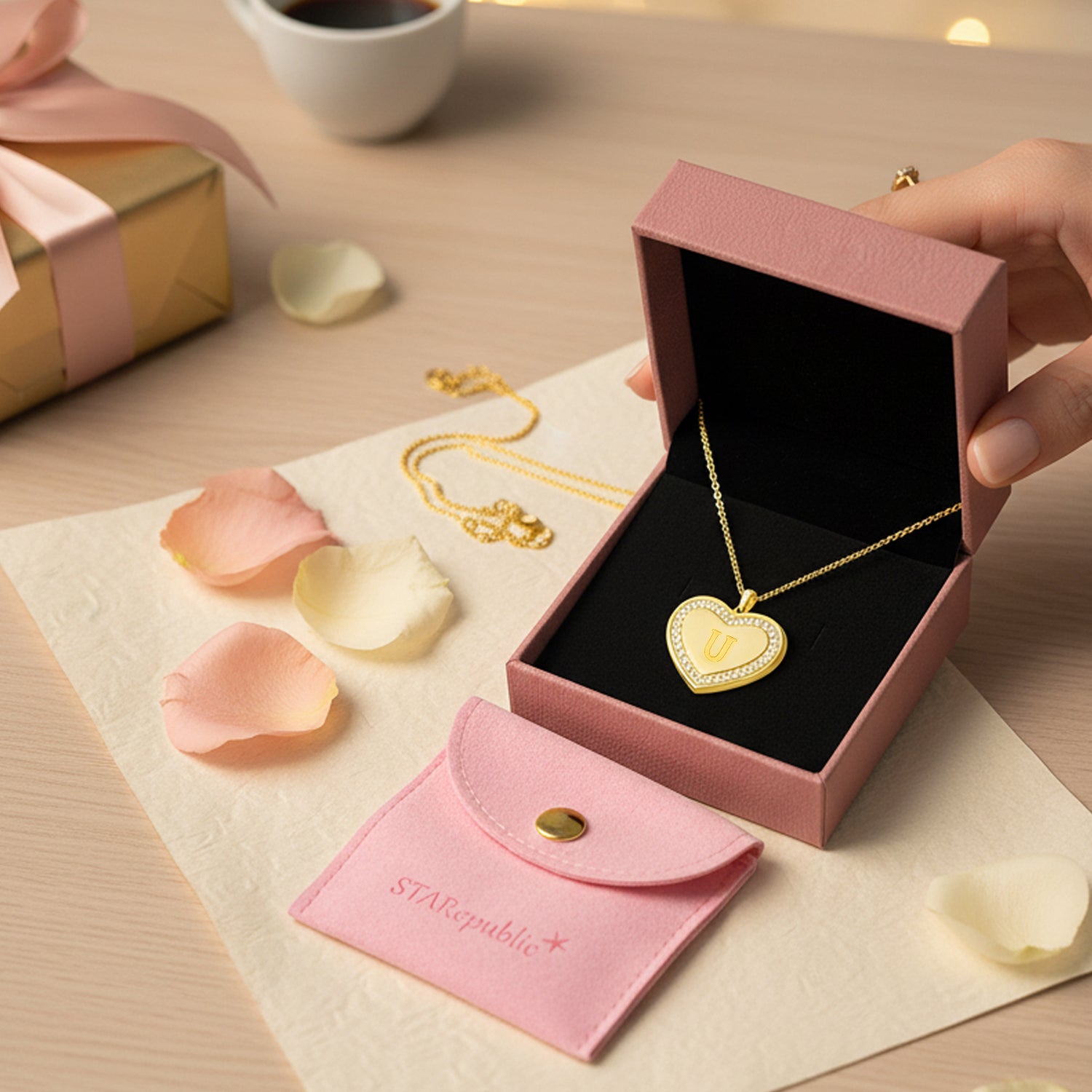 Gold heart-shaped necklace in a pink box with rose petals and a coffee cup in the background
